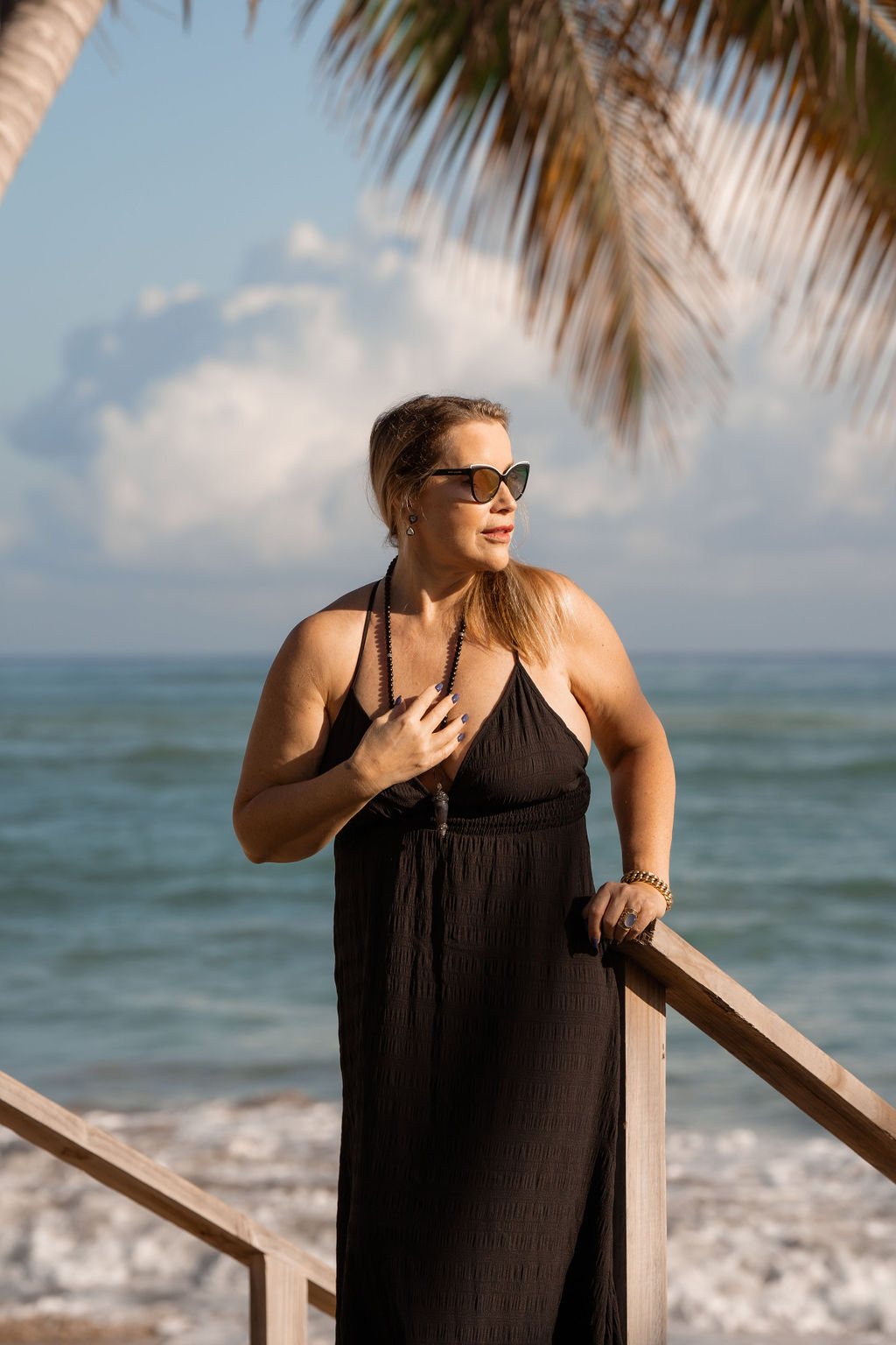 Juliette standing by the sea in a black dress, hand on a railing
